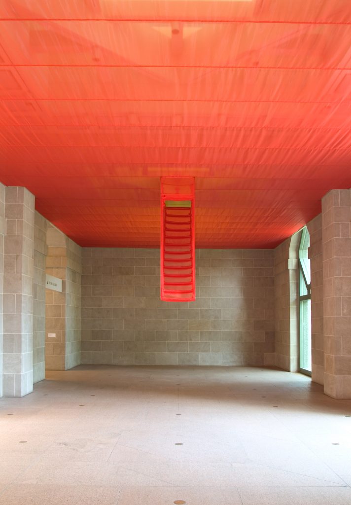 Red fabric culminating in the form of a partially descending staircase covers the ceiling of a large stone-walled gallery space.