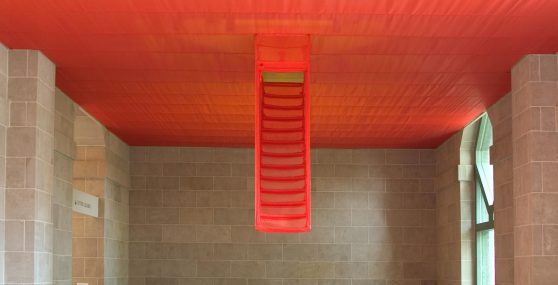 Red fabric culminating in the form of a partially descending staircase covers the ceiling of a large stone-walled gallery space.