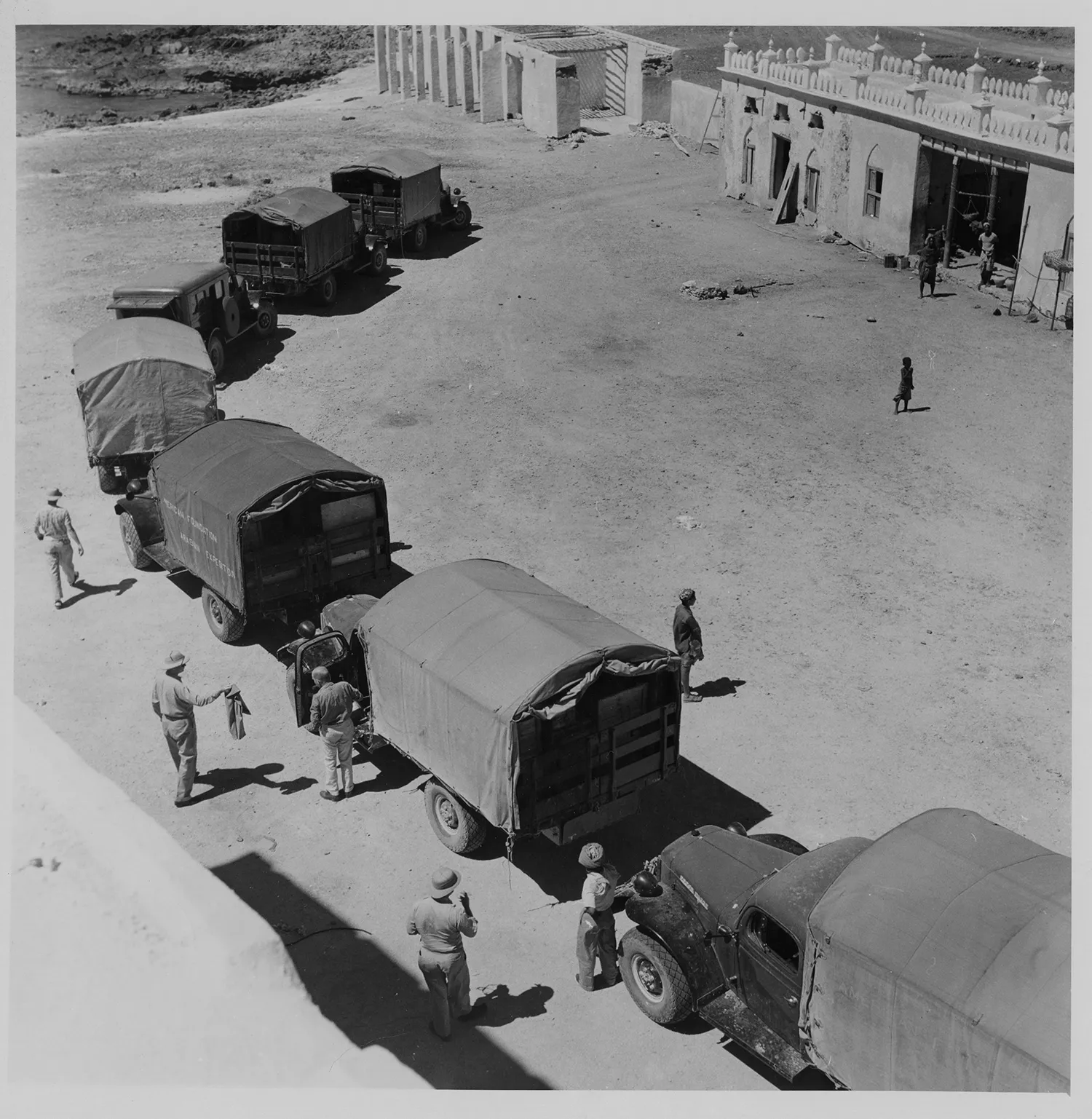 black and white photo - a curving line of trucks in front of a wall, some people walking nearby