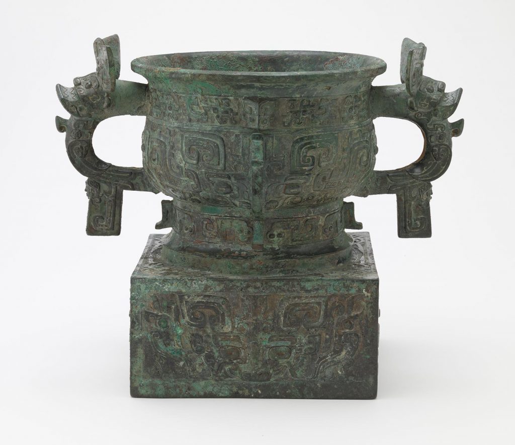 A patinated, incised bronze vessel with a handle on either side and a large rectangular base.