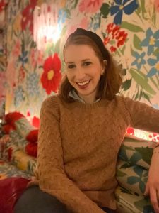 A portrait photo of a smiling woman with red hair pulled back with a headband and wearing a brown sweater.