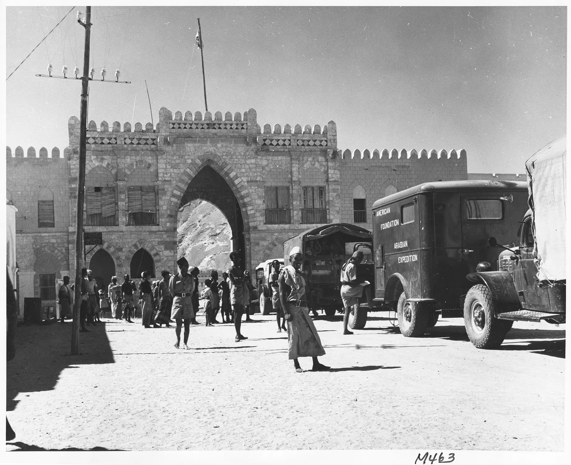 black and white photo with a queue of trucks approaching a wall with an arched opening, people walking nearby