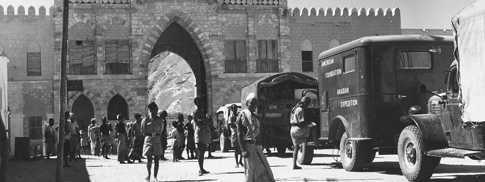 black and white photo with a queue of trucks approaching a wall with an arched opening, people walking nearby