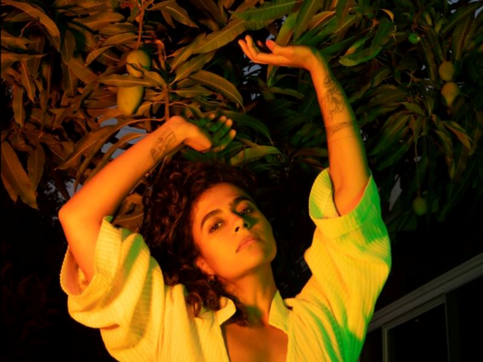 Photograph of woman from the chest up, arms lifted, and extending up into green leaf trees.