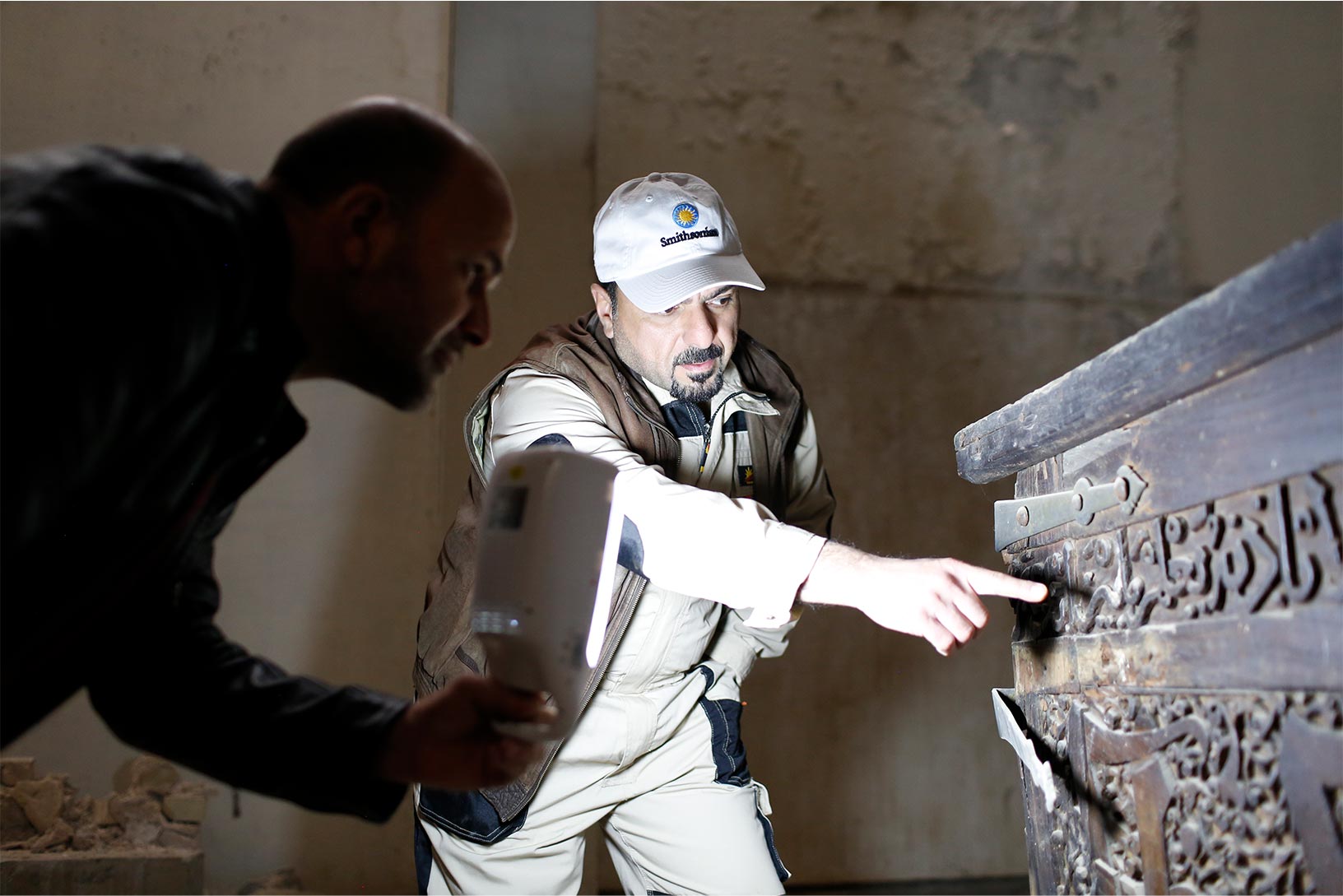 Two Smithsonian employees examine a worn chest in a dark building with stone walls. One employee shines a light while the other points to engraving on the chest.