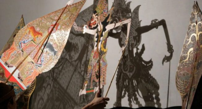 Shadow puppet made of intricately cut paper being controlled by puppeteer.