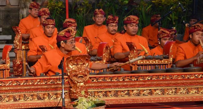 Orange-clad gamelan ensemble playing in front of a leafy background.