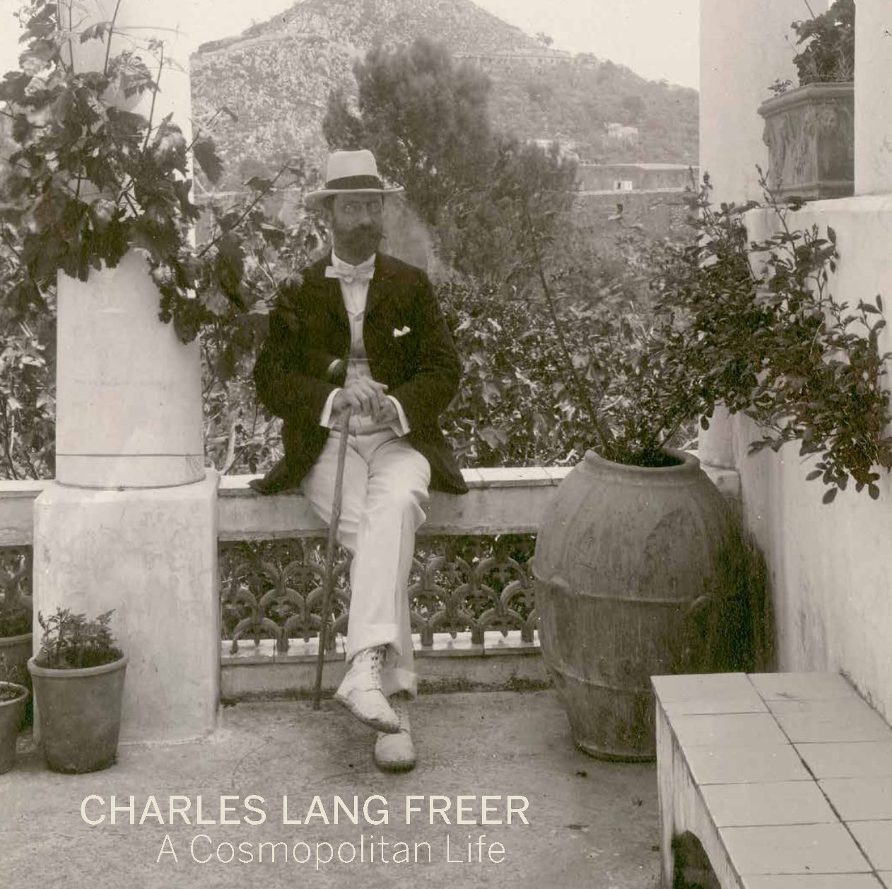 Charles Lang Freer: A Cosmopolitan Life book cover. A black and white photograph of Freer sitting on the edge of a balcony or porch, legs crossed, hands resting on a cane. He wears a dark suit jacket over light-colored shoes, pants, and shirt. Atop his head is a light-colored fedora. He is surrounded by houseplants. Behind him, in the distance, a large mountain covered with vegetation and small buildings rises up.