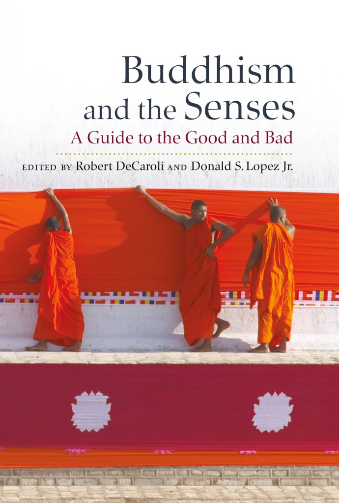 Buddhism and the Senses book cover. Three Buddhist monks wearing bright vermillion robes stand in front of a white wall holding up a large red cloth against the wall.