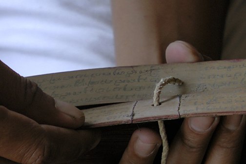 Hands holding two leaves of a lontar manuscript, bound with rough string.