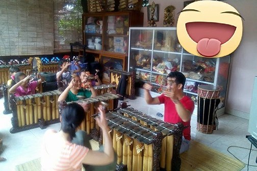 A man instructing a group of girls playing gamelan, with a large smiling emoji sticker in the top right quarter of the photo.