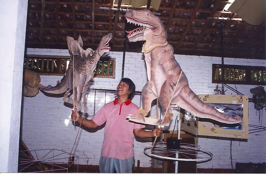 A man holding two large, ornate dinosaurs puppets, a t-rex and stegosaurus.