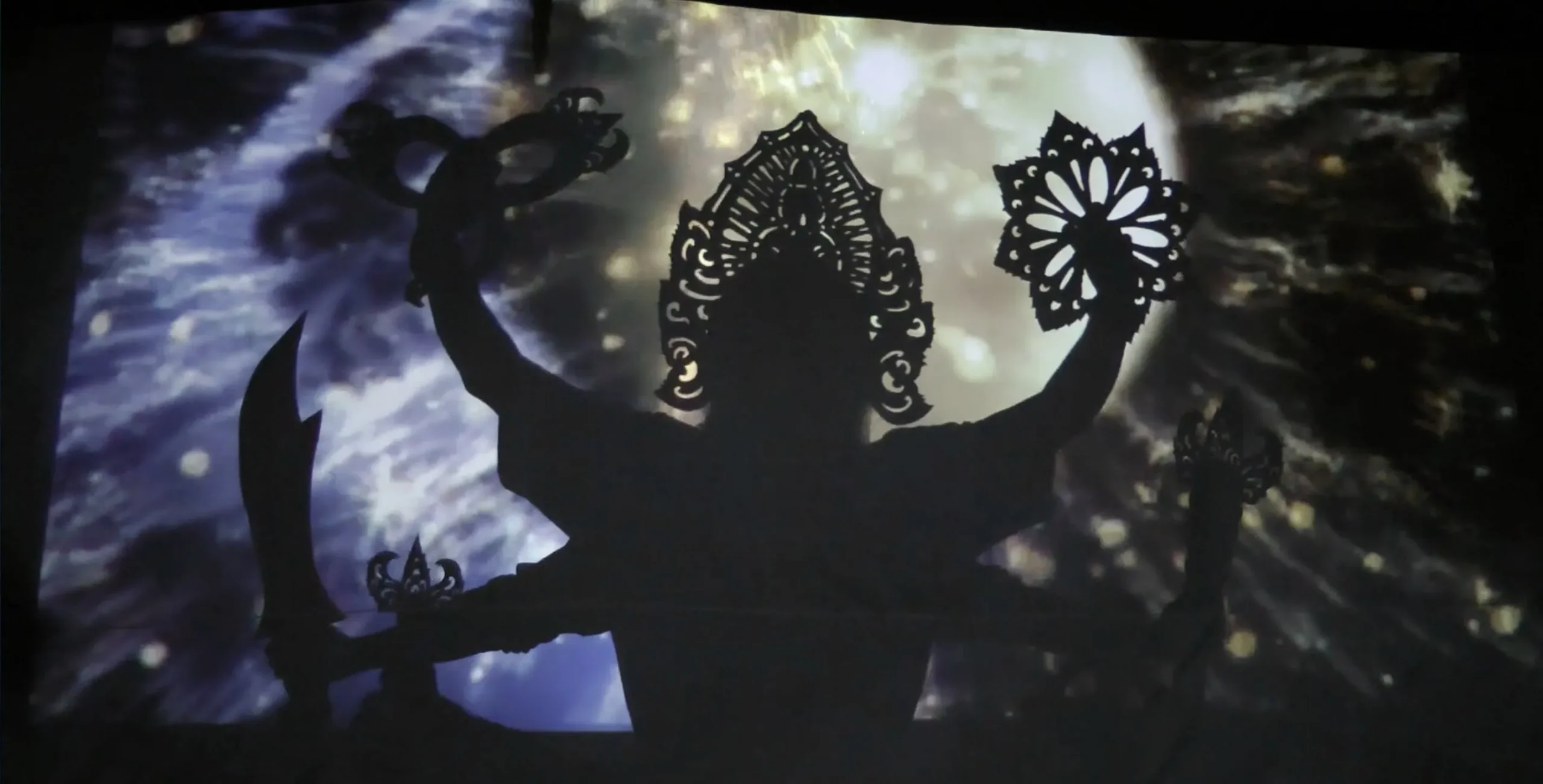 the silhouette of a six armed figure, in front of an abstract projected background.