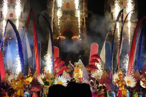 Ornately costumed dancers in front of a backdrop of red, blue, and white flags, with fireworks shooting into the air.