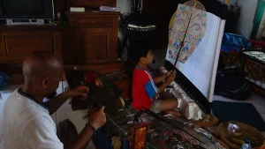 A man practicing gender and his young son holding a leaf-shaped Kayon puppet, inside a home.