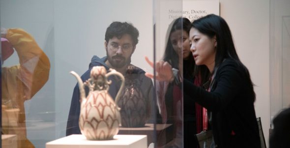 A woman points at a spouted ceramic vessel in a case, speaking to a man and a woman nearby.