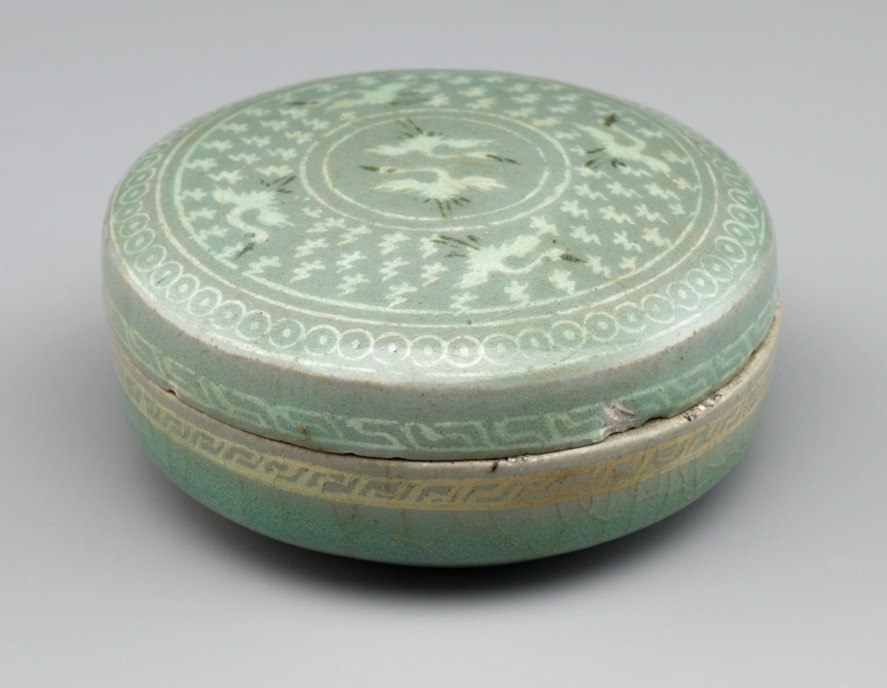 A lidded, green ceramic vessel with white decoration of birds and geometric patterns on top.