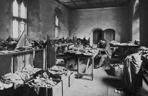 Black and white photo of a man, head resting in his hand, thinking, at a table amidst other tables covered in books and scraps of materials.