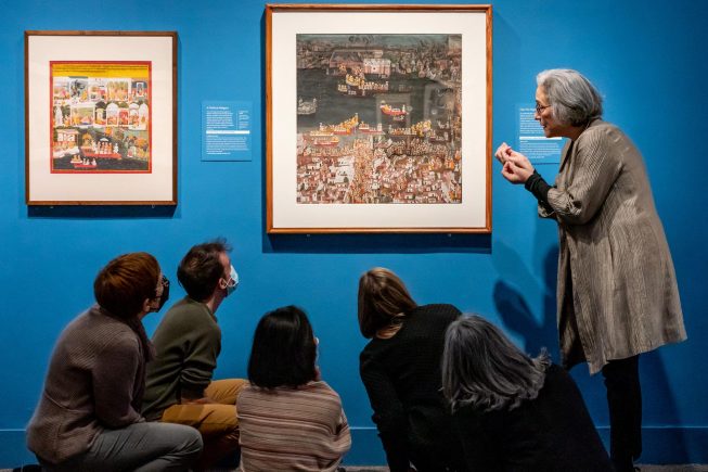 Five people kneel below two South Asian paintings. A woman stands on the right, gestures at the rightmost painting, and speaks.