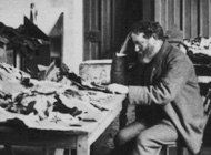 Black and white photo of a man, head resting in his hand, thinking, at a table amidst other tables covered in books and scraps of materials.