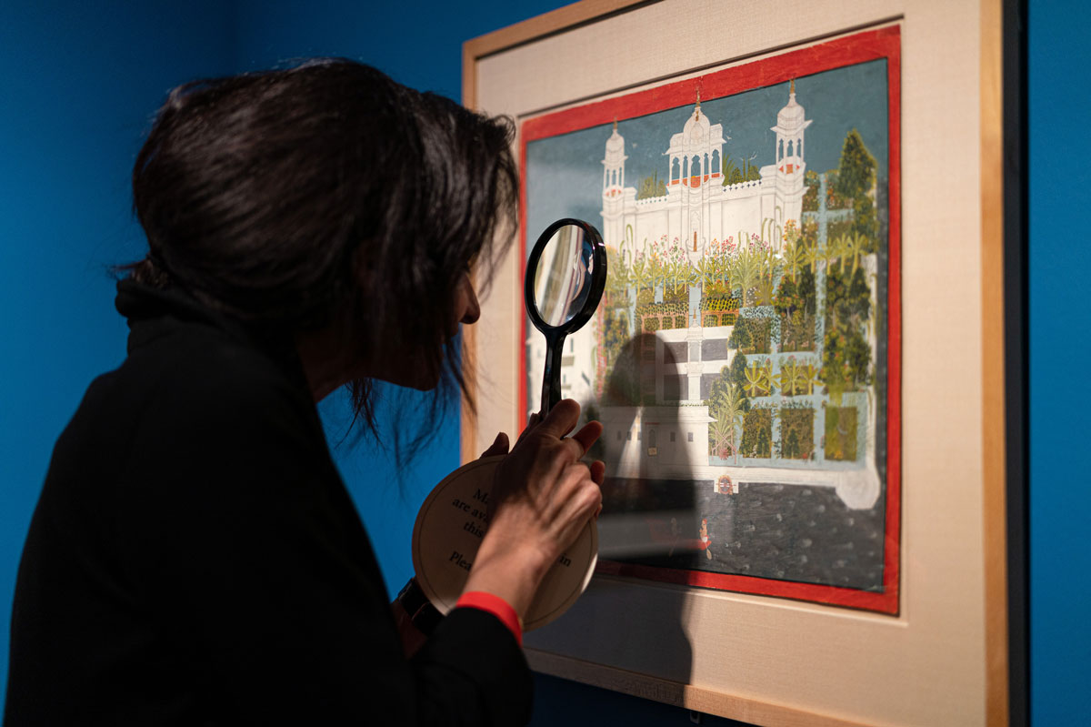 A woman holds up a magnifying glass before a detailed painting of a palace, framed against a deep blue wall.