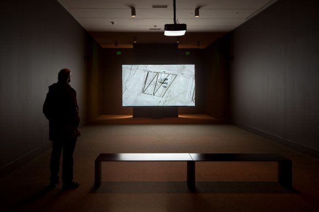 photo of a darkened gallery with a figure watching a projected overhead image of a structure