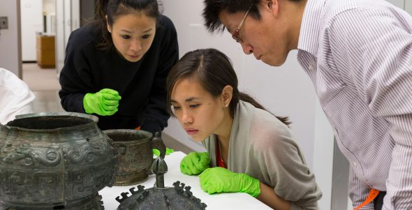 Three individuals look closely at two bronze vessels and a lid