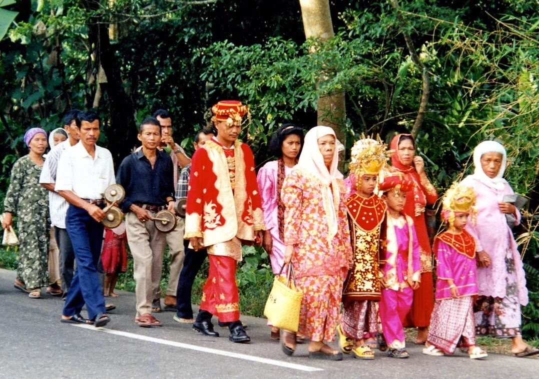 Wedding procession lead by individuals in colorful traditional garments in red, gold, and pink.