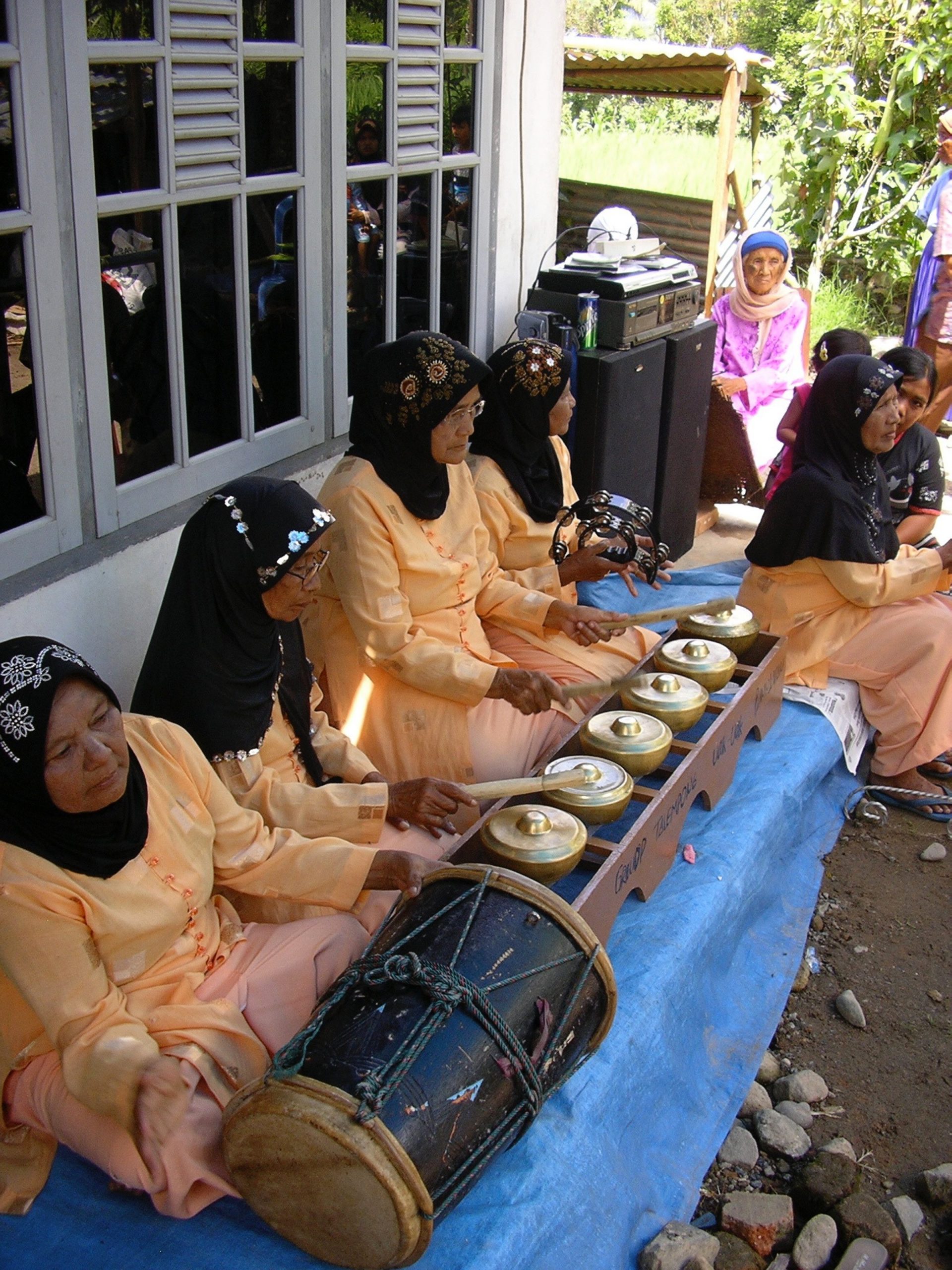 Women in orange garments and black head coverings perform talempong outside in the shade of a house.