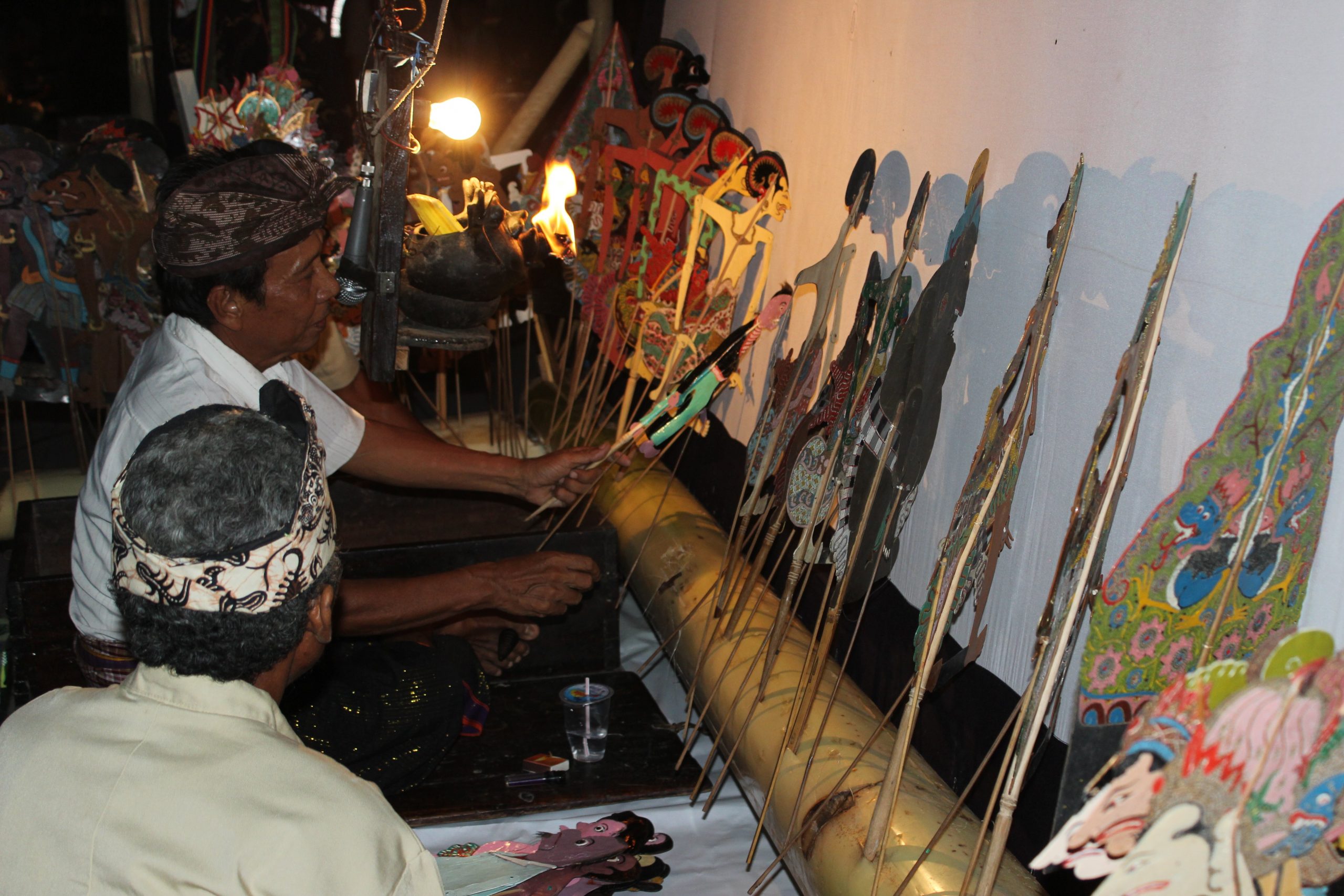 A puppeteer selects a wayang puppet from row of puppets lined up against a wall.