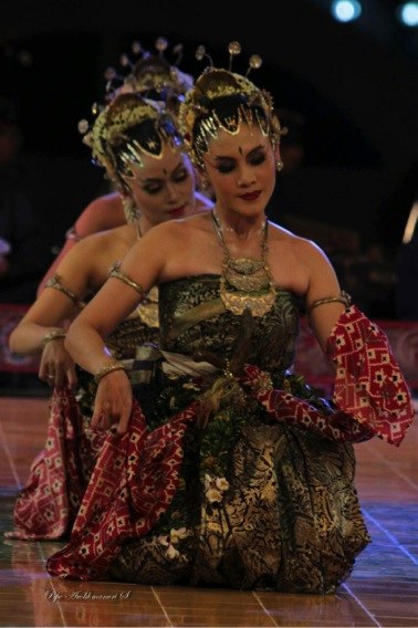 A female dancer in ornate costume, with two matching dancers kneeling in the same pose behind her.
