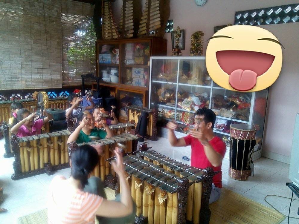 A man instructing a group of girls playing gamelan, with a large smiling emoji sticker in the top right quarter of the photo.