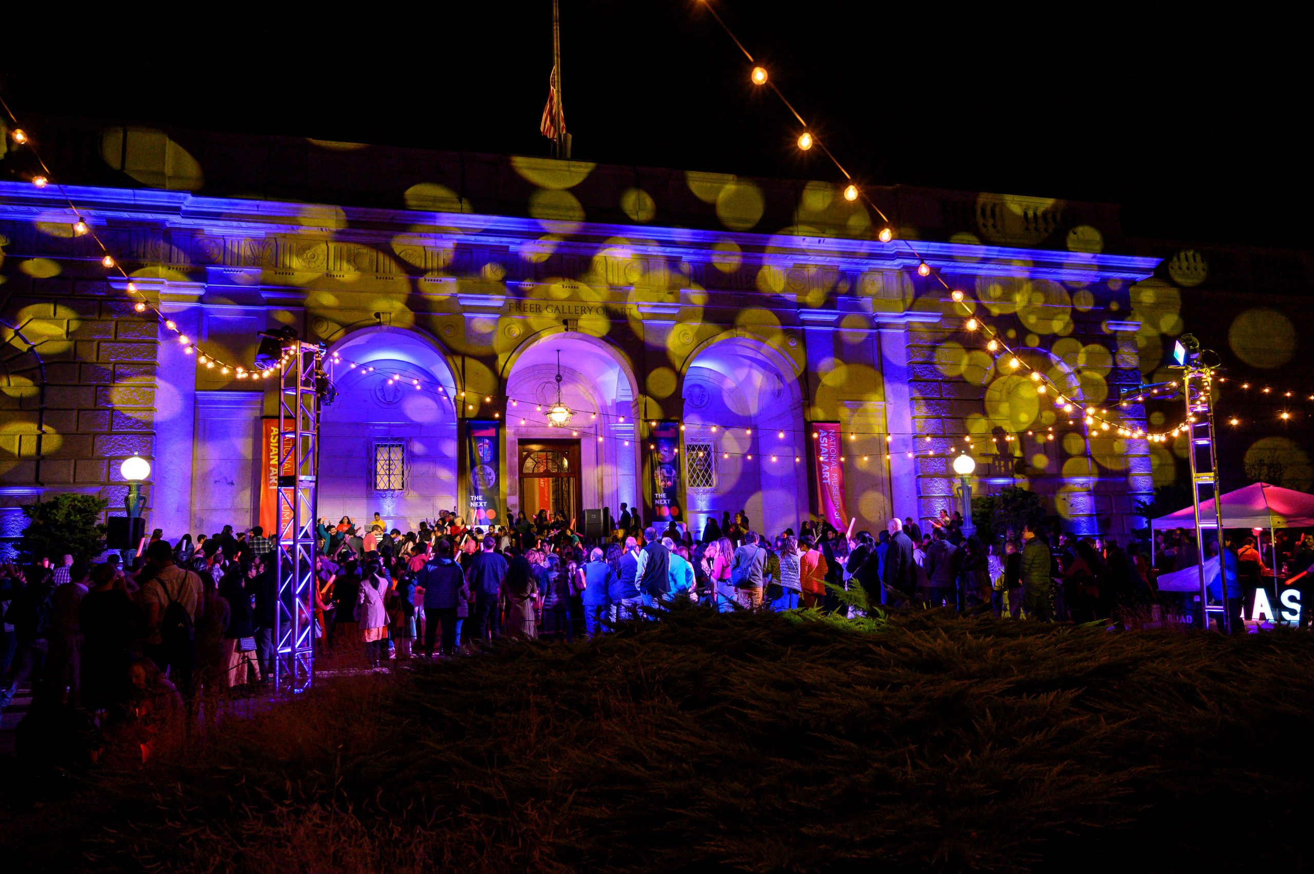 A building facade at nighttime with colorful lights projected on the building and people gathered on the plaza out front.