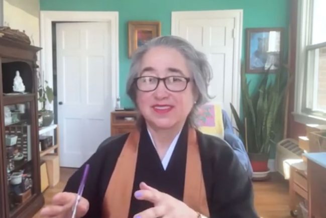 Woman with short grey hair and glasses sitting in a room speaking on camera