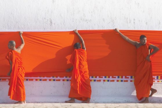 Four people with shaved head and simple orange robes hold up an orange banner across a white wall.