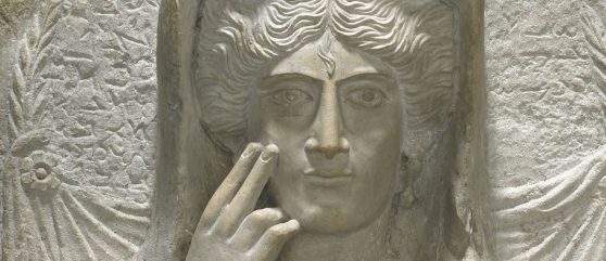 detail from a sculpture of a woman showing her touching her cheek with two fingers, made of white stone.