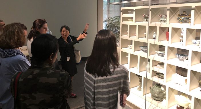 A docent shows a tour group a shelf full of ceramics.