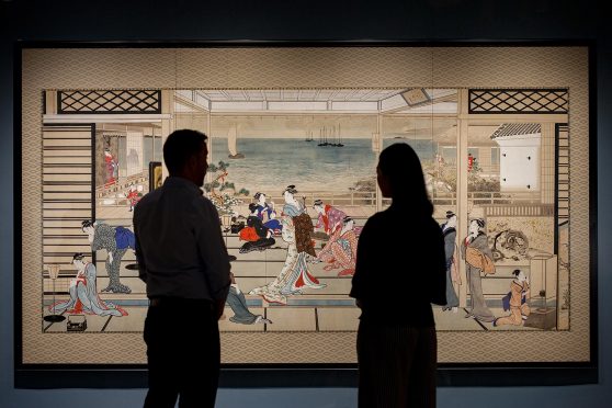 Two people stand in silhouette before a wide woodblock print of a large room filled with women in kimonos.
