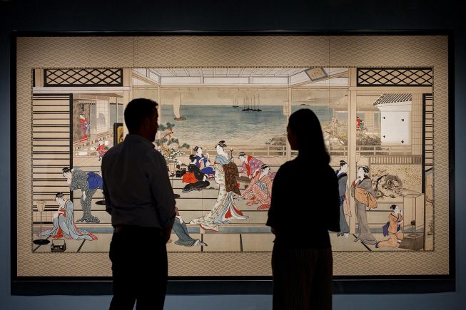 Two people stand in silhouette before a wide woodblock print of a large room filled with women in kimonos.