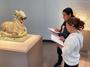 A statue of a kneeling cow sits on a pedestal. To its left, two people hold pencils over sketchbooks.