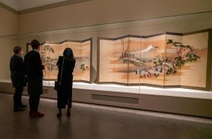 Three people stand close together and look at two large folded screens, both painted with Japanese landscapes.