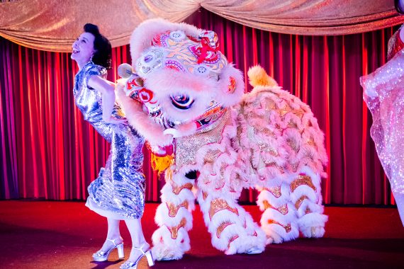 A woman in a silver dress dances on stage with performers in a lion-dance costume.
