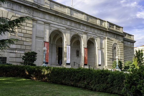 The stone, arched building facade of the Freer Gallery of art seen from an angle against a blue sky with white clouds. In the foreground is a grassy area with a line of green bushes