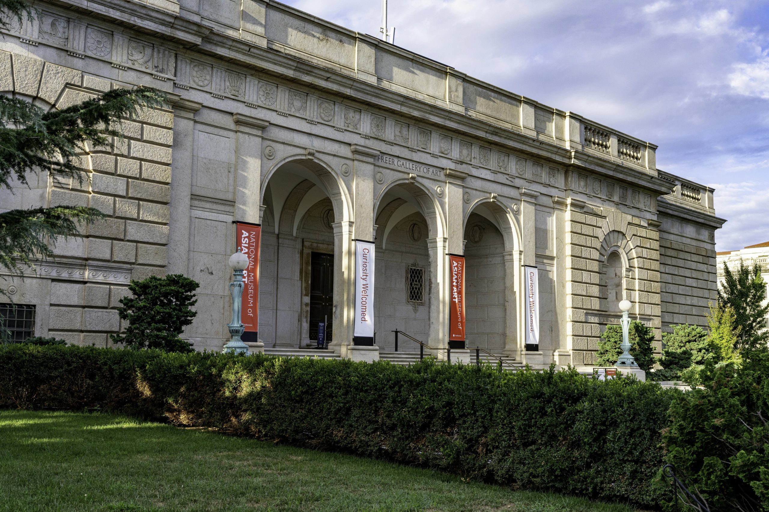The stone, arched building facade of the Freer Gallery of art seen from an angle against a blue sky with white clouds. In the foreground is a grassy area with a line of green bushes