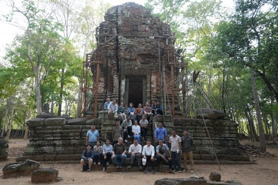 A large group of researchers sit on the stairs of a stone temple at Koh Ker