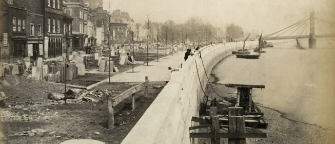 Sepia-tone photo of a walkway along a river with buildings under construction.
