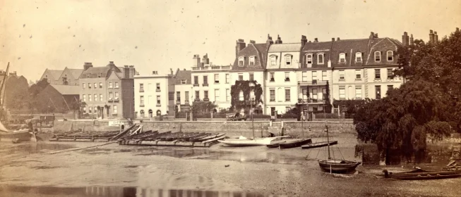 Sepia-tone photo of a boatyard before a row of buildings in London.