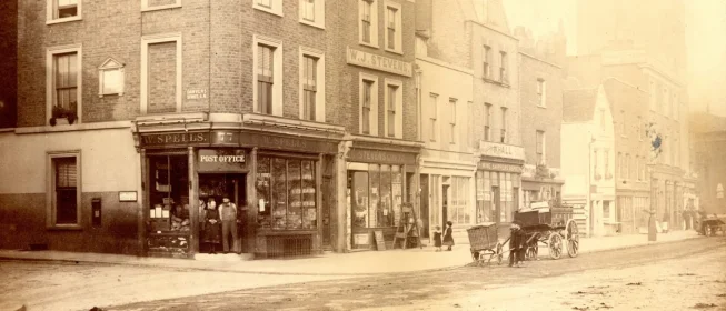 Sepia-tone photo of a L London street corner showing a post office and a building ensigned, W.J. Stevens. A horse-drawn carriage is parked out front.
