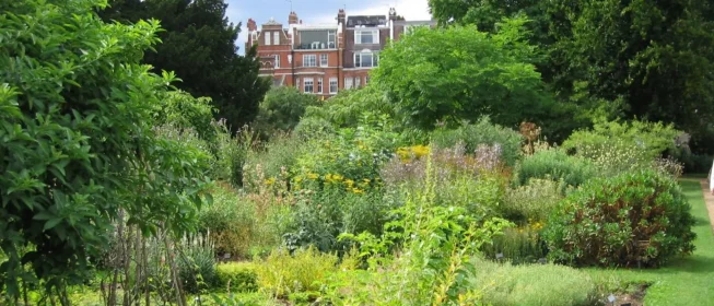 A lush garden, with a lavish home visible in the background, framed by copses of trees