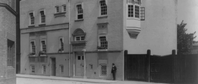 Black and white photo of a large London home with a man standing in front, leaning against the facade.